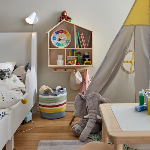 Un lit extensible BUSUNGE blanc à côté d’une tente pour enfants et d’une table pour enfants, plus une maison de poupée/étagère murale sur le mur.