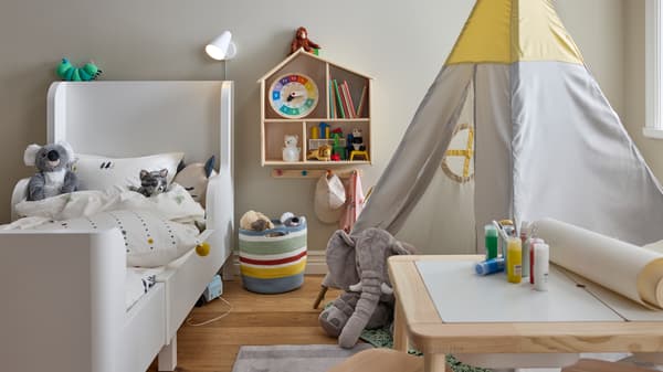 Un lit extensible BUSUNGE blanc à côté d’une tente pour enfants et d’une table pour enfants, plus une maison de poupée/étagère murale sur le mur.