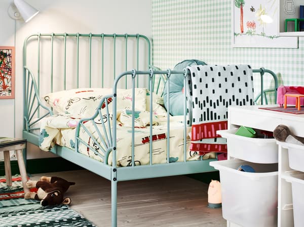 Un lit extensible MINNEN turquoise dans l’angle d’une chambre pour enfants, avec des jouets, une lampe de bureau et des rangements.