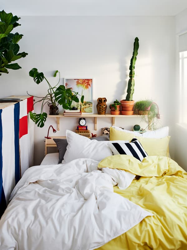 Un lit NEIDEN avec deux parures de lit ÄNGSLILJA, une jaune et une blanche. Des plantes et des décorations sur une étagère au-dessus du lit.