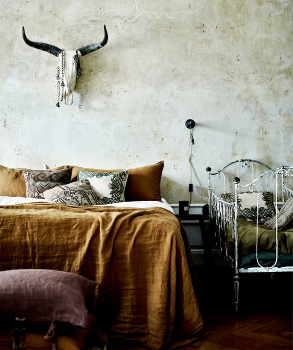 Un lit recouvert de draps couleur rouille surplombé d'un crâne d'animal fixé au mur et à côté d'un berceau en métal blanc.
