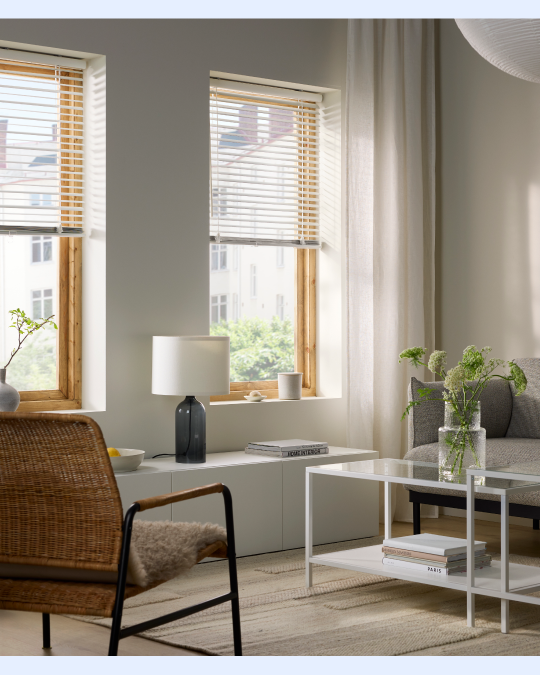 Un living con una mesa de centro de vidrio y en grente un sillón de mimbre. Las ventanas tiene cortinas venecianas.