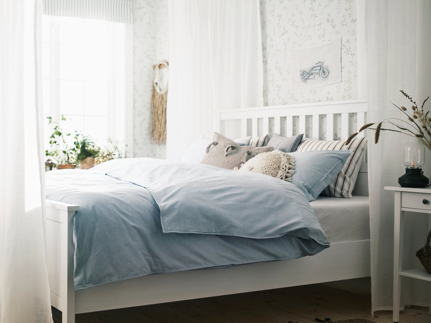 Un llit HEMNES blanc en una habitació plena de llum que està envoltat de cortinetes SILVERLÖNN de color blanc, amb coixins i roba de llit blava al damunt.
