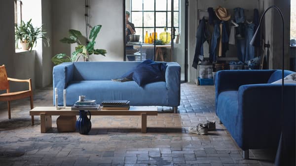 Un loft de style industriel avec sur un sol en pierre beige une table basse rectangulaire longue en bois avec autour deux KLIPPAN avec une housse en jean.