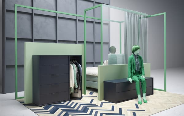 Un mannequin vêtu de vert assis sur une commode basse, à côté d’une commode plus haute avec des tiroirs et des vêtements qui y sont suspendus.
