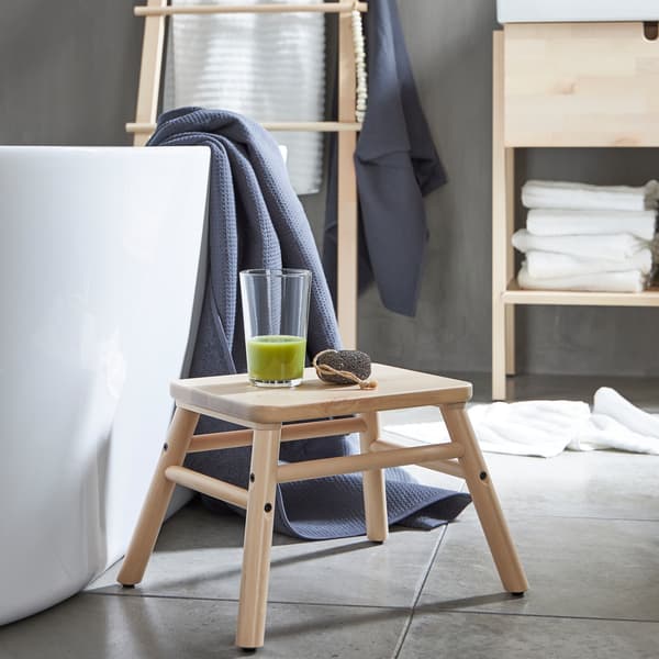 Un marchepied VILTO en bouleau à côté d’une baignoire où est posé un verre de smoothie vert, dans une salle de bain minimaliste.