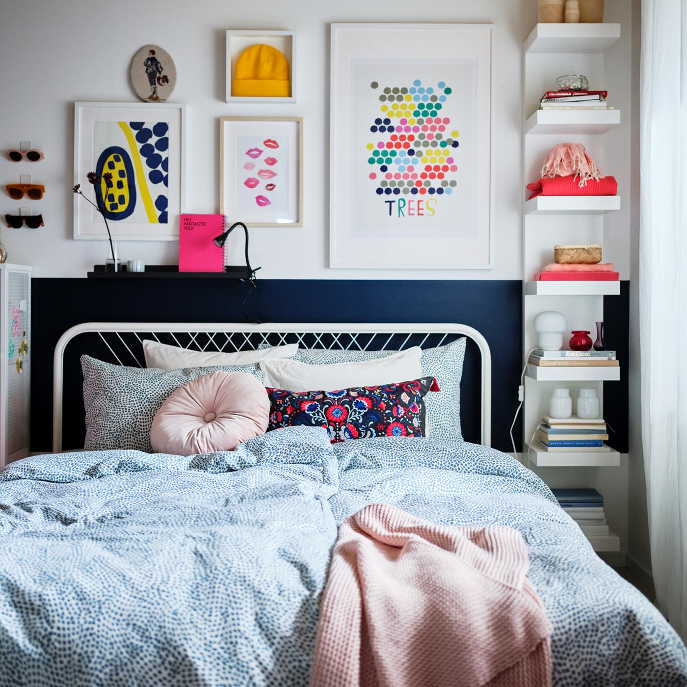 Un marco de cama NESTTUN blanco con una funda nórdica TRÄDKRASSULA blanca/azul, con arte colorido y estantes abiertos en la pared de atrás.