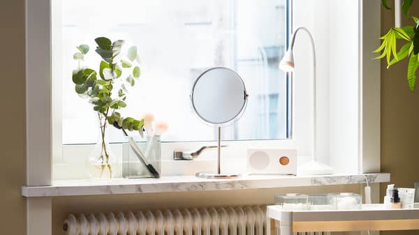 Un miroir est près d'un rebord de fenêtre à côté d'une lampe de table blanche, d'un brin vert dans un vase et d"un haut-parleur Bluetooth sur le rebord de la fenêtre.
