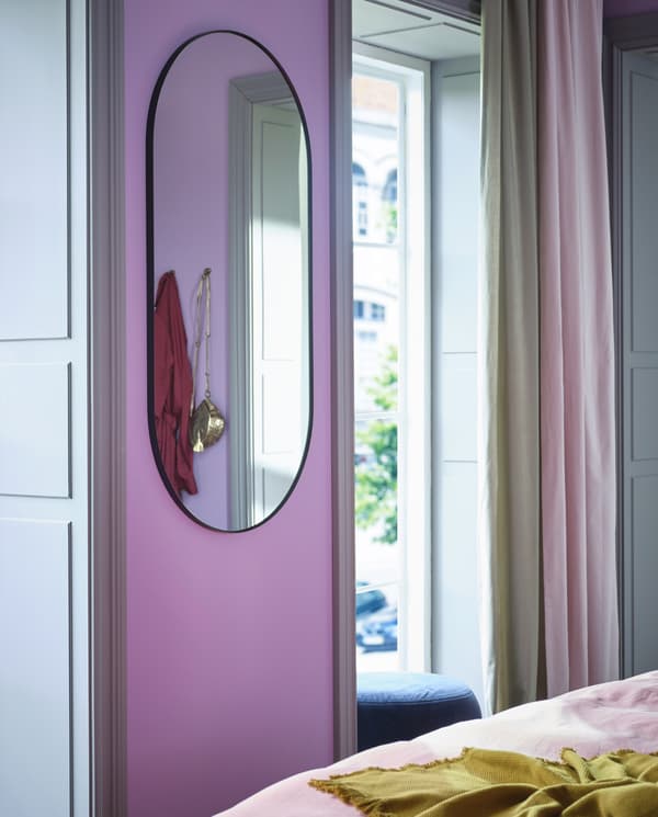 Un miroir ovale LINDBYN à cadre noir, sur un mur étroit entre deux fenêtres décorées de rideaux en beige et rose.