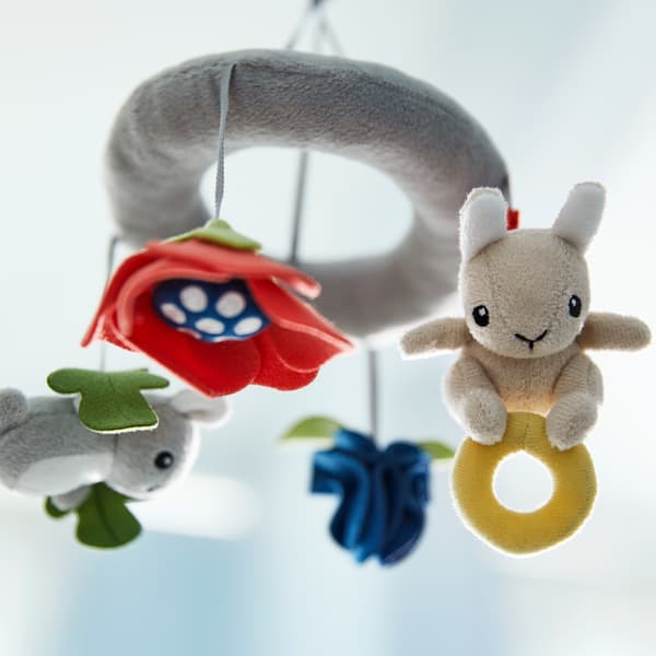 Un móbil GULLIGAST de varias cores pendurado lixeiramente en ángulo nun cuarto luminoso cun coelliño de peluche diante.