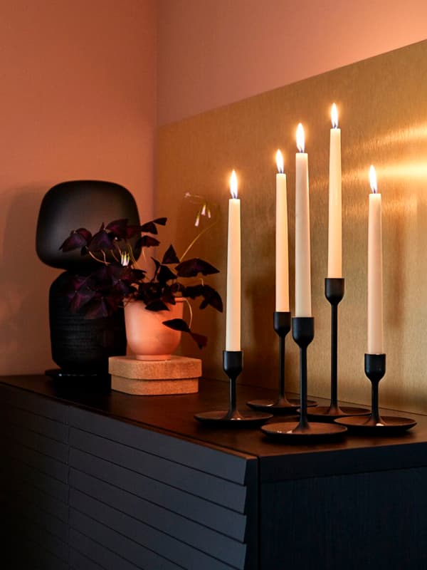 Un mobile con sopra 5 candielieri con candele accese FULLTALIG. Accanto, un vaso con una pianta e una lampada con cassa.