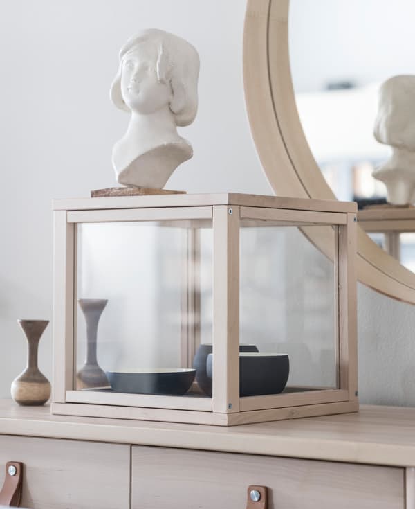 Un mobile con una teca in vetro SAMMANHANG di IKEA. Al suo interno, alcune ciotole e sopra un busto di una statua.