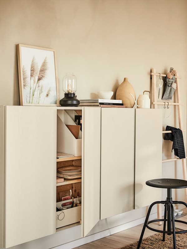 Un mobile dalle ante beige appoggiato alla parete di cui una aperta che svela materiali da disegno e quderni. Davanti al mobile uno sgabello nero. 