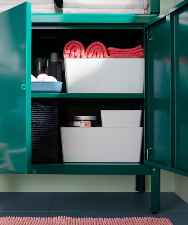 Un mobile di metallo verde con ante semiaperte, pieno di cestini e contenitori con gli accessori per il bagno - IKEA