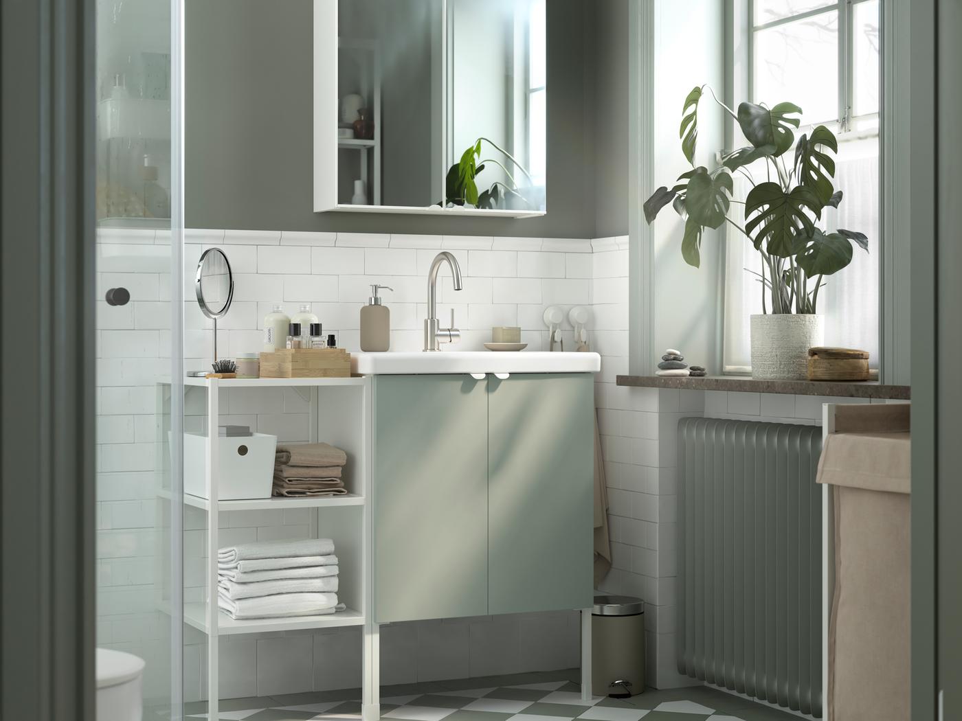 Un mobile ENHET bianco con due ante e un ripiano a giorno a sinistra riempito con asciugamani in un bagno grigio e bianco.