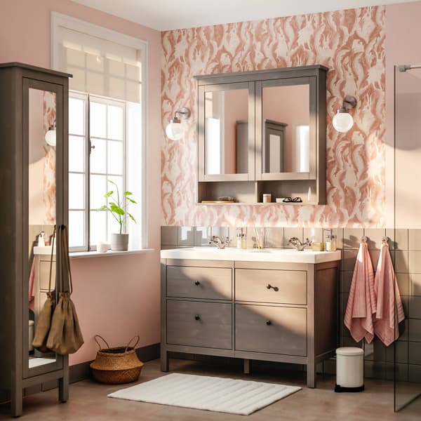 Un mobile grigio da bagno con lavabo. Sopra, due specchi con a lato due lampade a parete. Le pareti sono sui toni del rosa. A lato, c'è una finestra ampia da cui entra luce.