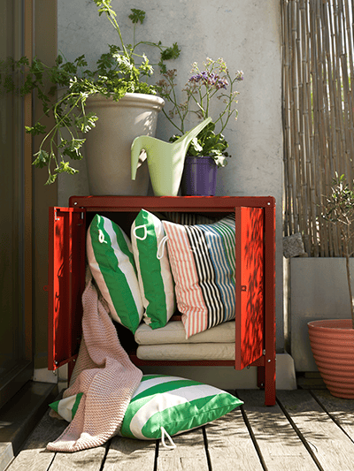 Un mobile rosso con le ante aperte che contiene vari cuscini su un balcone. Sopra, un vaso con una pianta e un annaffiatotio IKEA PS 2002.