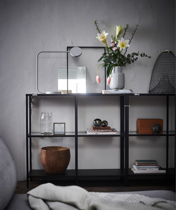 Un módulo de exposición aberto en fronte dunha parede gris, cun arranxo floral, libros e elementos de decoración.