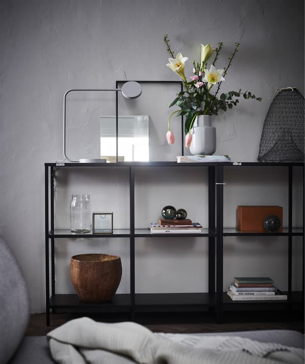 Un módulo de exposición abierto en frente de una pared gris, con un arreglo floral, libros y elementos de decoración.