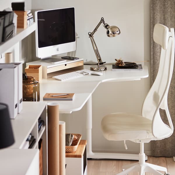 Un monitor en un soporte para monitor sobre un escritorio de esquina BEKANT blanco con una silla de oficina en blanco/beige, y un estante de pared LACK blanco.