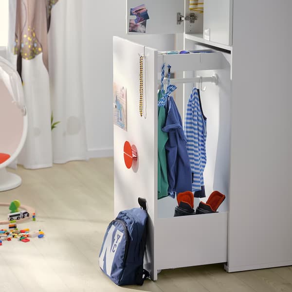 Un mueble SMASTAD blanco con un módulo extraíble abierto para colgar y guardar ropa infantil, situado a la altura de los niños. Se ve también una mochila azul apoyada en el frente del mueble. 