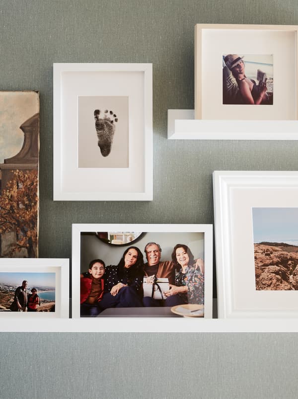 Un mur avec deux tablettes pour photos blanches et plusieurs cadres RIBBA blancs avec des photos de la famille et des vacances, et l’empreinte du pied d’un bébé.