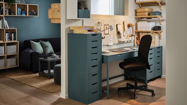 Un mur sépare un salon et un espace de travail à la maison. Bureau et caisson à tiroirs gris-turquoise, chaise de bureau noire.