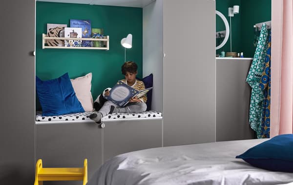 Un neno sentado cun libro nun canto de lectura con coxíns, unha lámpada e un andel de libros, dentro dunha parede de módulos de almacenaxe en gris.