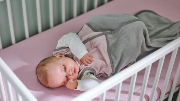 Un neonato in un sacco nanna LEN rosa dorme in un lettino GULLIVER bianco su un materasso rivestito da un lenzuolo con angoli LEN rosa - IKEA.