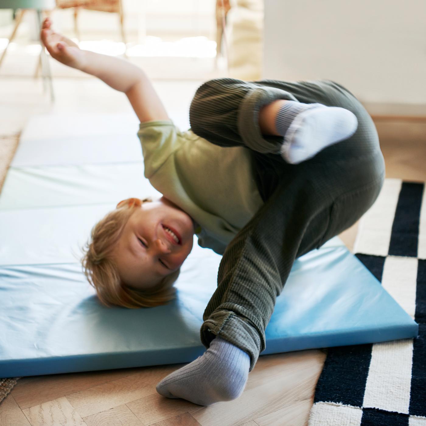 Un niño con pantalones verde oscuro y jersey verde claro hace una voltereta en una colchoneta plegable PLUFSIG azul.