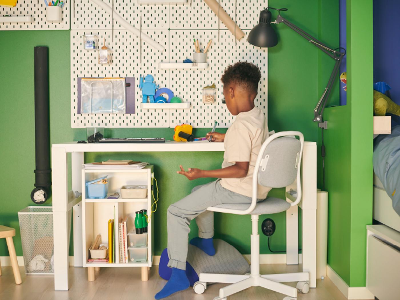 Un niño en una silla de escritorio para niños ÖRFJÄLL en un escritorio PÅHL blanco, junto a una combinación de tableros perforados SKÅDIS blancos en una pared verde.