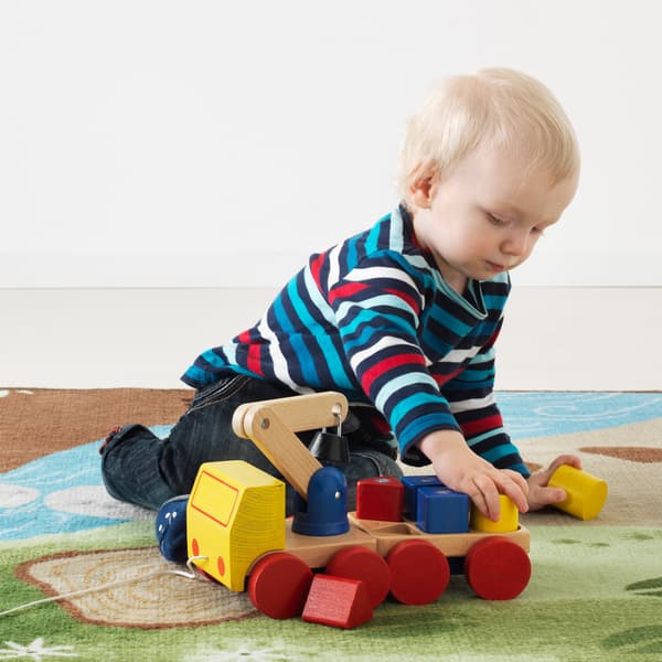 Un niño está jugando con una grúa de madera roja, azul y amarilla IKEA MULA con bloques sobre una alfombra infantil en verde y azul.