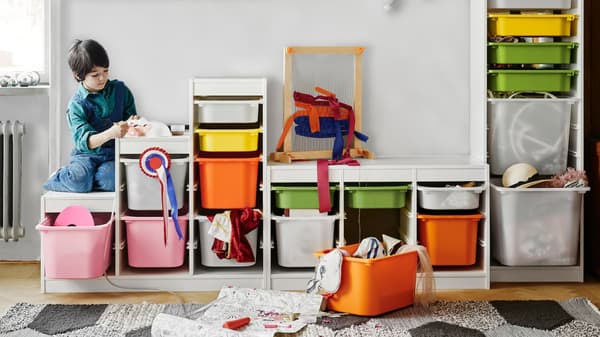 Un niño sentado sobre un mueble TROFAST de distintas alturas con cajas plásticas, grandes y pequeñas, de distintos colores. Una de las cajas está fuera del mueble y contiene artículos para hacer manualidades. 