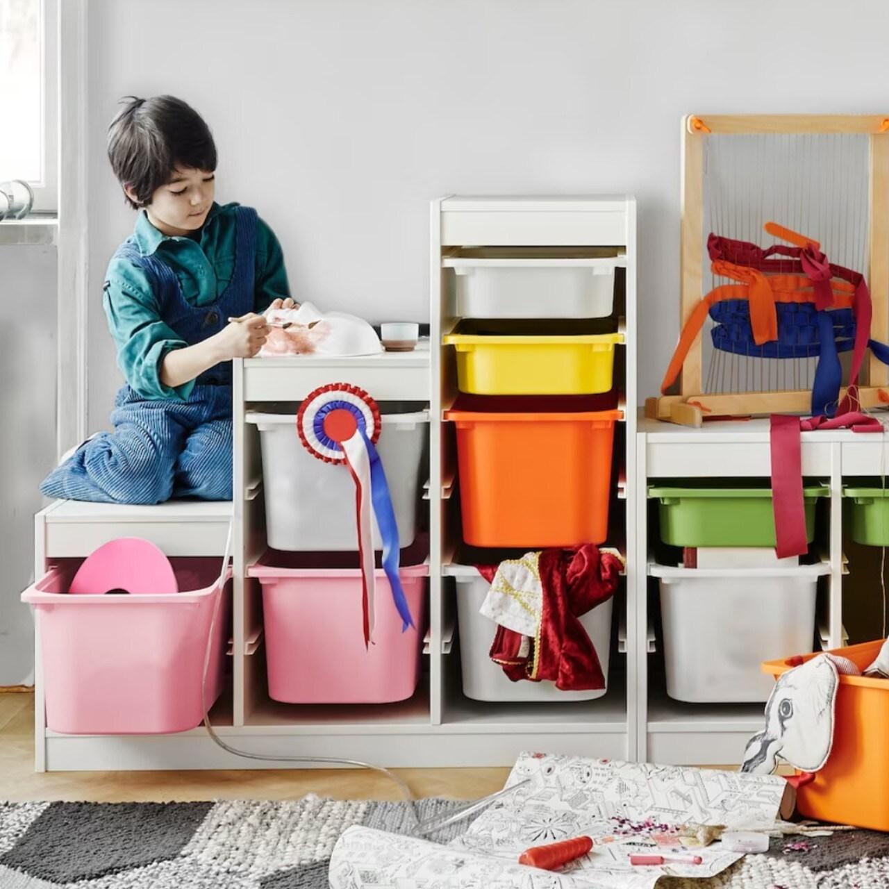 Un niño sentado sobre un mueble TROFAST de distintas alturas con cajas plásticas, grandes y pequeñas, de distintos colores. Una de las cajas está fuera del mueble y contiene artículos para hacer manualidades. 