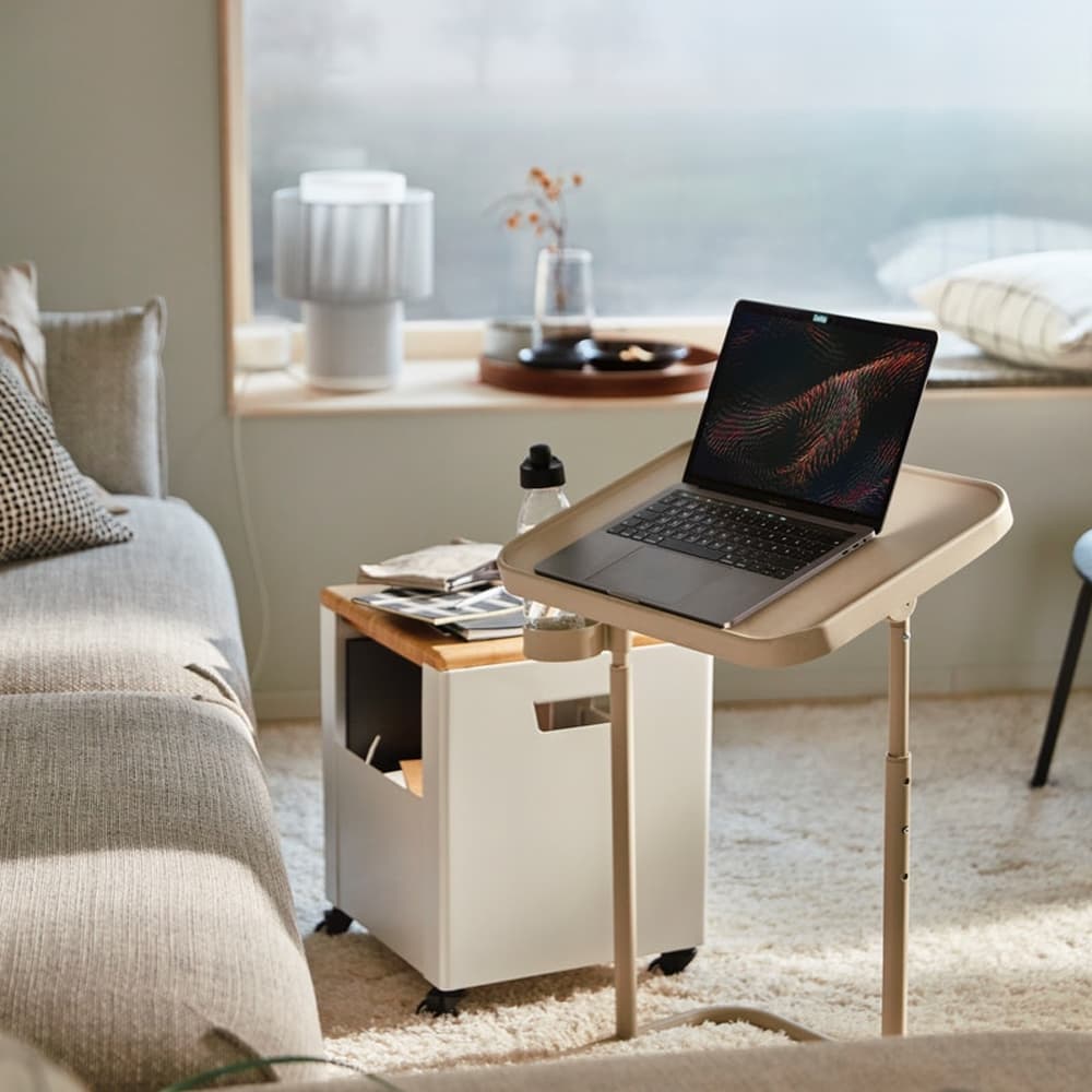 Un ordinateur posé sur une table d'appoint inclinée devant un canapé en tissu beige et une table basse à roulettes.
