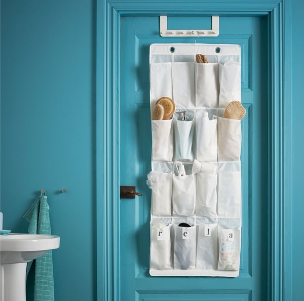 Un organizator pentru pantofi SKUBB, plin cu diverse accesorii pentru baie, agățat de o ușă, într-un decor de baie turcoaz.