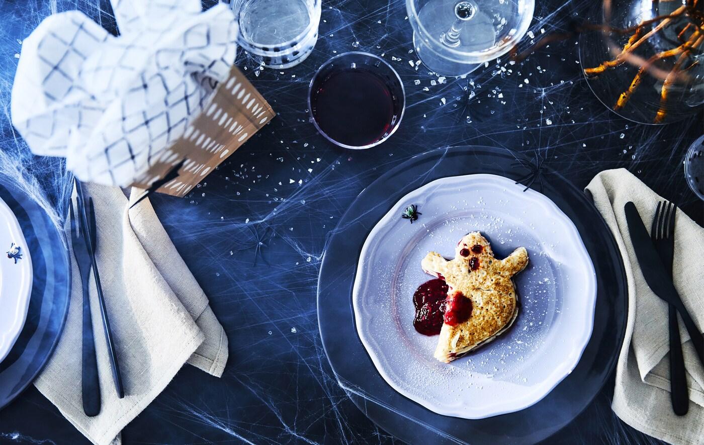 Un pancake a forma di fantasma con confettura che ricrea l'effetto sangue, su un piatto bianco circondato da stoviglie e un regalo incartato.