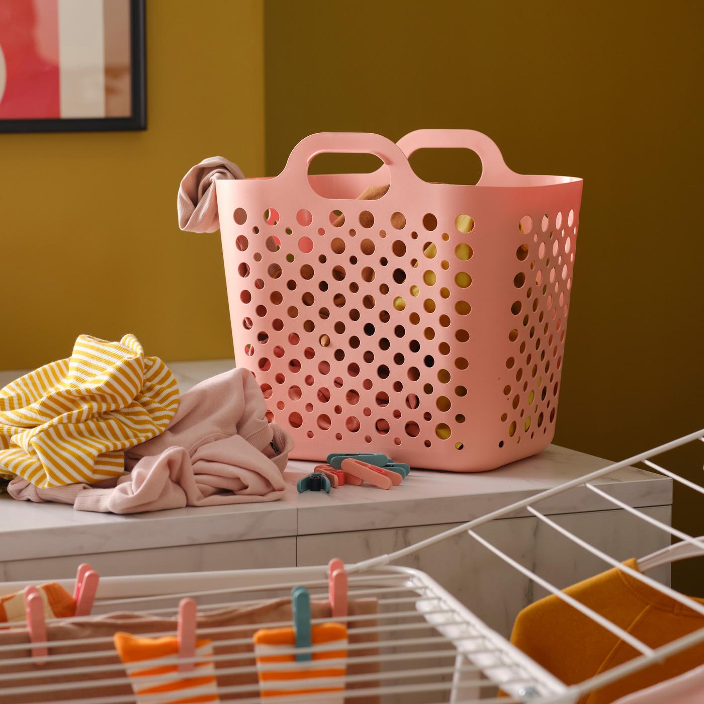 Un panier à linge souple SLIBB rose et une pile de vêtements sur un plan de travail de cuisine, plus un séchoir blanc avec d’autres vêtements.