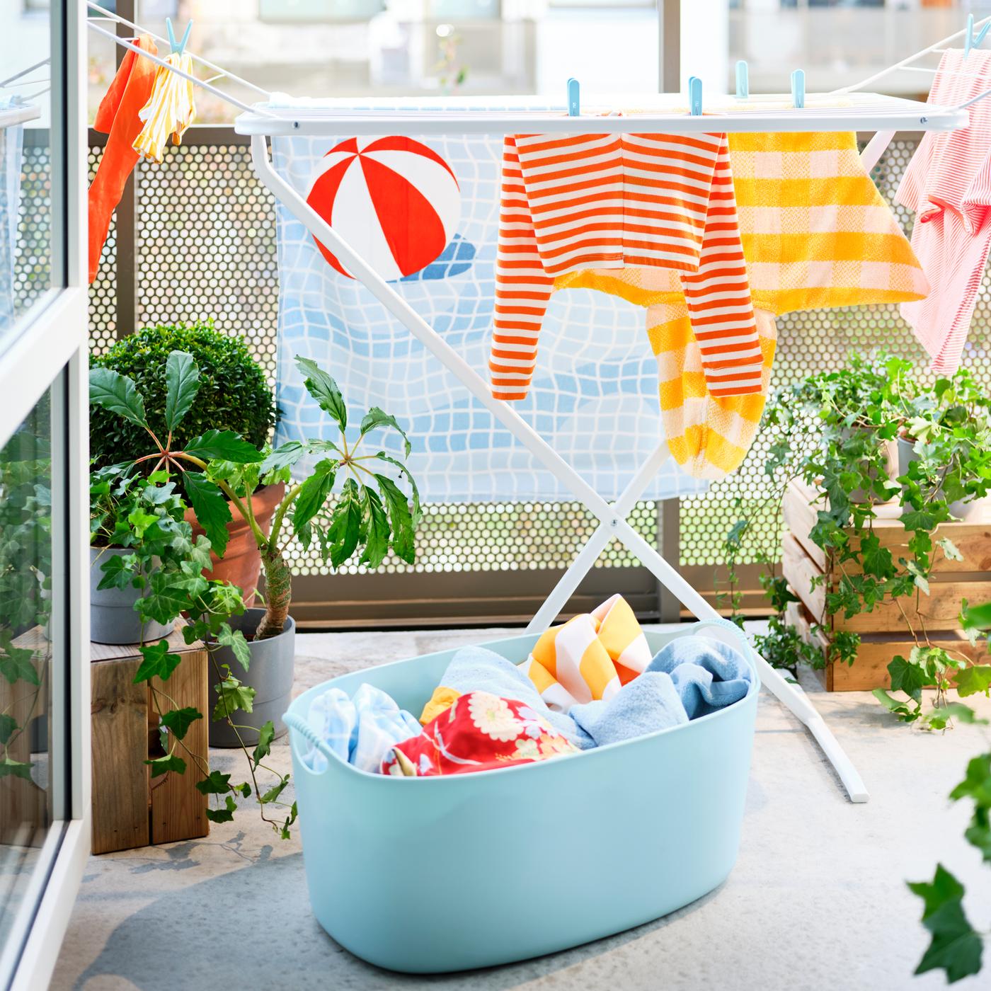 Un panier à linge souple TORKIS bleu avec du linge dedans et un séchoir blanc sur un balcon ensoleillé, décoré avec des plantes.