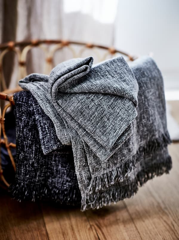 Un panier en rotin tressé brun clair sur un sol en bois contenant des plaids INGRUN pliés et débordant sur le côté.