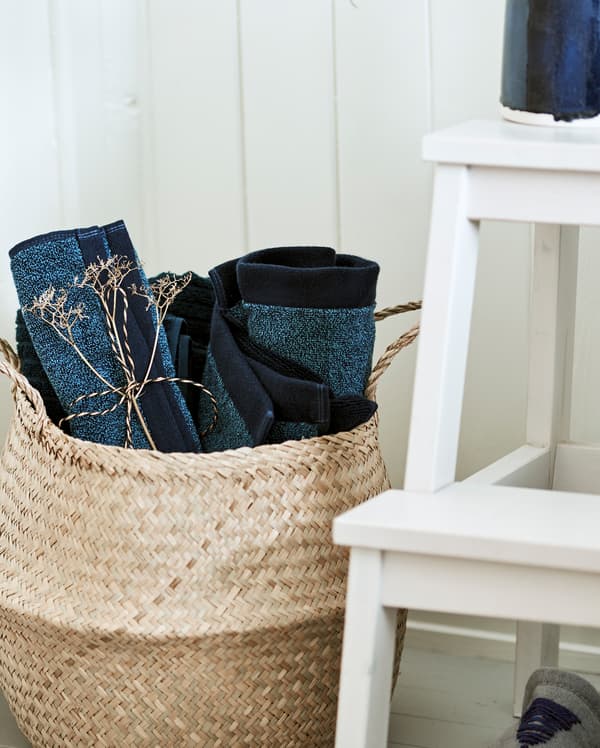 Un panier réalisé en un matériau tissé dans lequel sont enroulées des serviettes bleues, placé à côté d’un escabeau blanc.