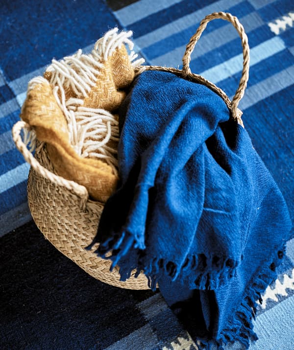 Un panier tissé contenant un plaid jaune à franges et un plaid bleu en lin sur un tapis aux motifs rayés bleu.