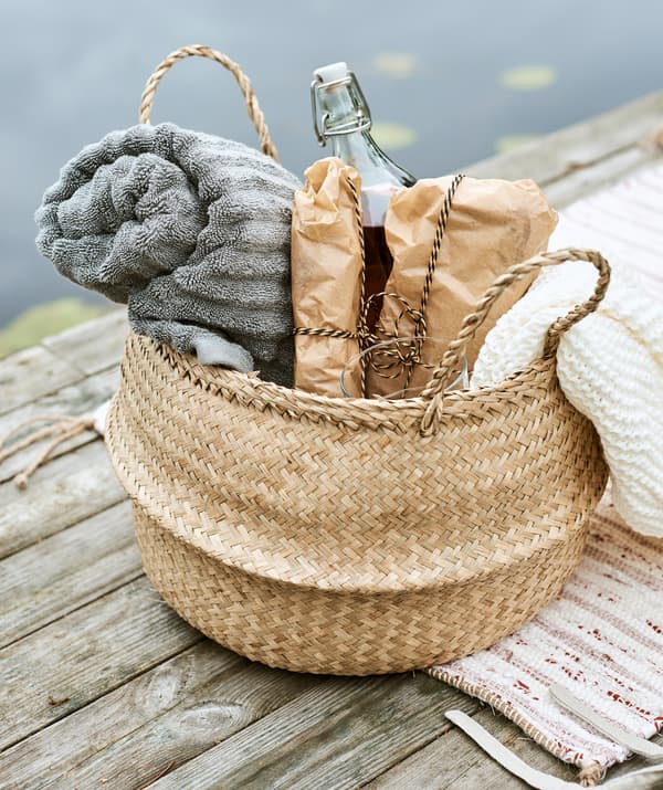 Un panier tressé prêt pour un pique-nique dans lequel se trouvent une serviette grise, des sandwiches emballés dans du papier, un plaid couleur crème et une boisson dans une bouteille.