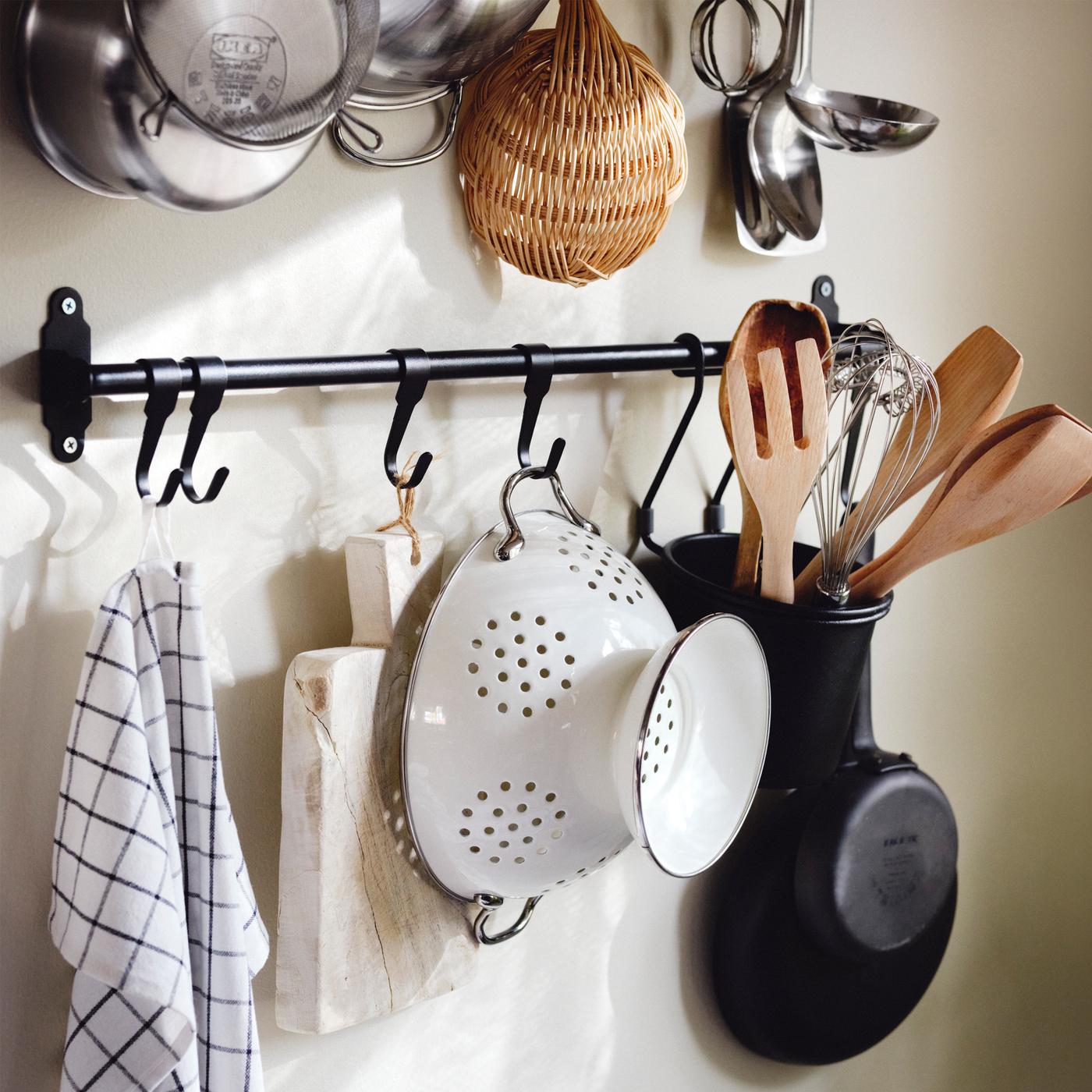 Un paño de cocina, una tabla de cortar, un colador blanco y utensilios en un recipiente HULTARP negro colgado de un riel HULTARP negro.