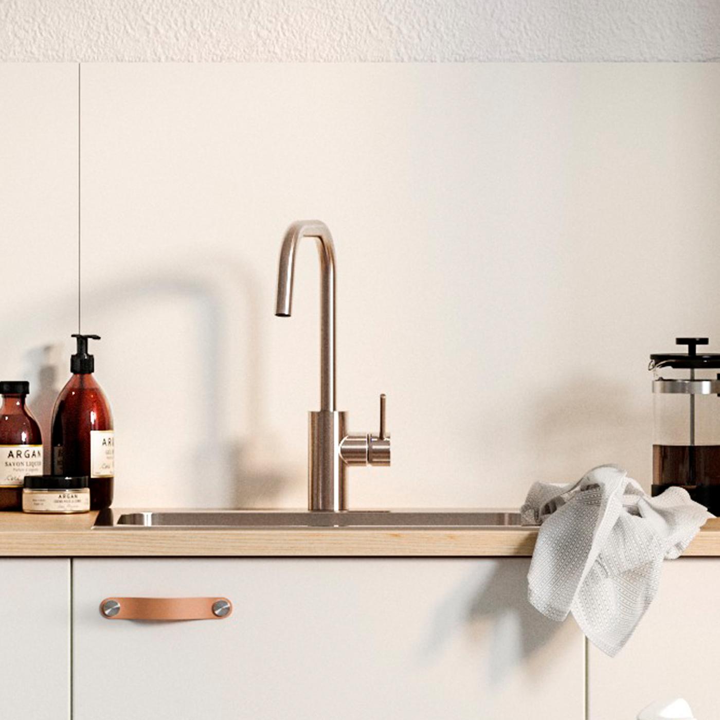 Un panou de perete alb, un front de bucătărie alb cu mânere din piele sub o chiuvetă, o cafetieră presă franceză și săpun cu aspect handmade.