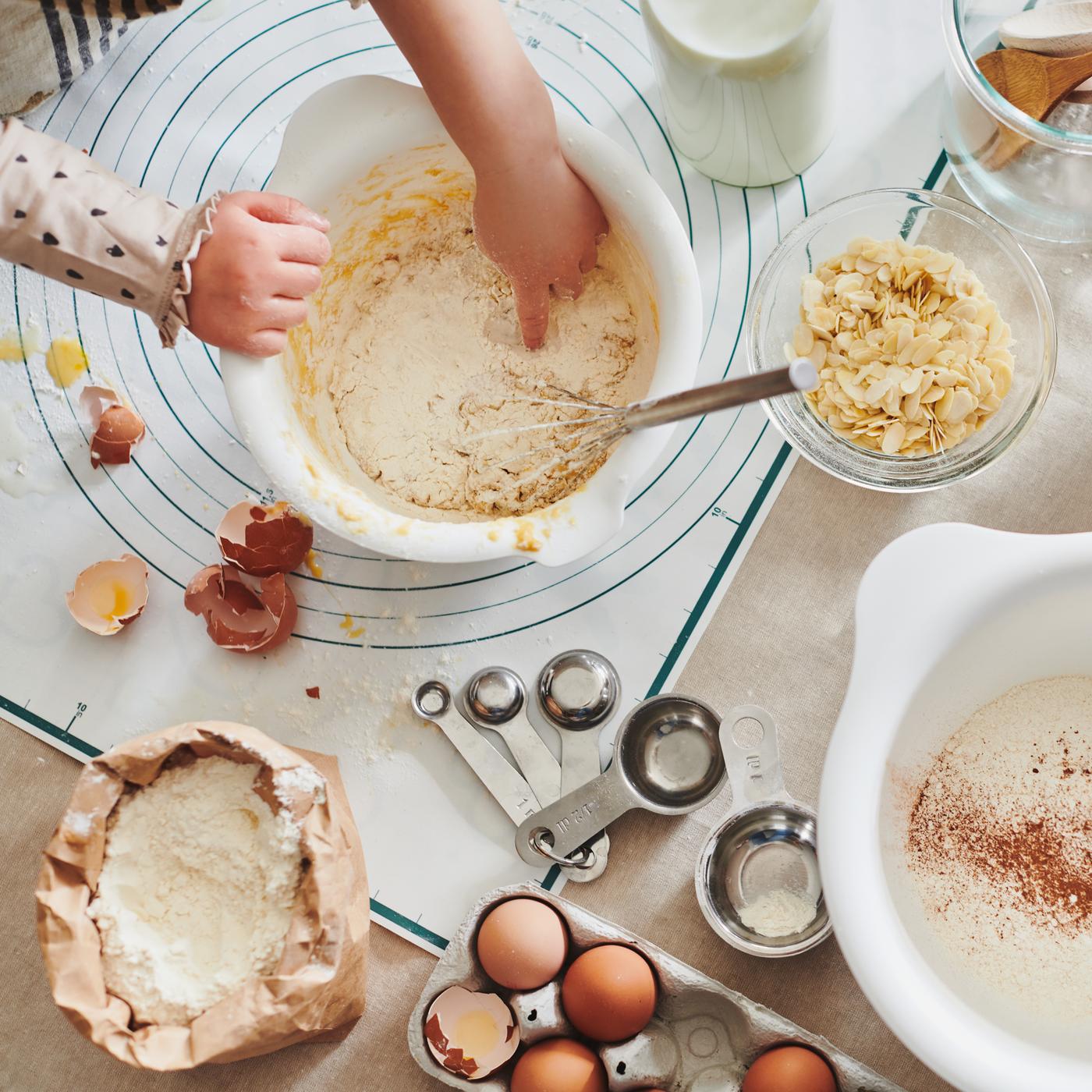 Un par de manos de niños trabajando la masa en un bol para mezclar blanco VISPAD sobre una mesa con huevos, harina y utensilios.
