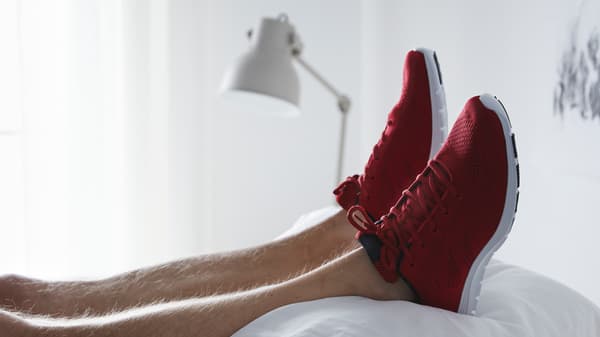 Un par de pies con zapatillas deportivas de color rojo brillante se encuentran encima de una pila de almohadas blancas y frescas, con una lámpara al fondo.
