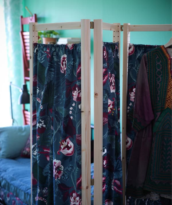 Un paravento con struttura in legno chiaro e tessuto floreale. Sullo sfondo sono appese ad una parete di color verde due sedie rosse FRÖSVI.