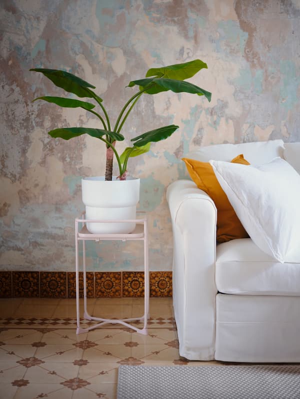 Un pedestal per a plantes de color rosa clar amb un test a sobre, al costat d'un sofà blanc amb coixins.