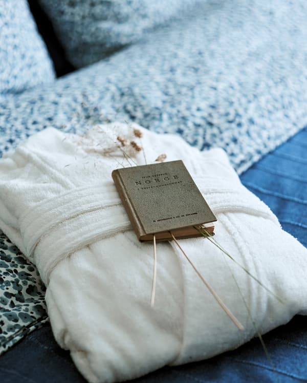 Un peignoir tout doux blanc plié sur un lit avec des couvertures bleues. Un vieux livre sur la Norvège renfermant quelques tiges d’herbe est posé sur le lit.